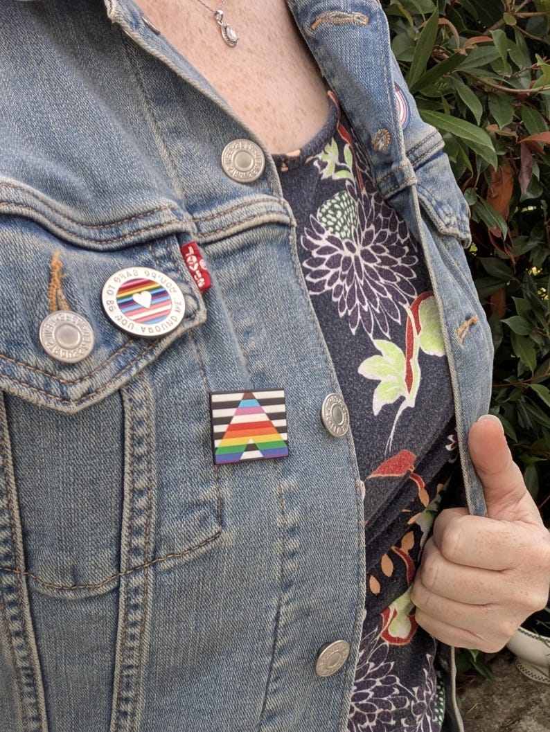 Peut inclure: Veste en jean bleu clair avec deux épingles. L'une a un motif de cœur arc-en-ciel et l'autre un motif de triangle noir, blanc et arc-en-ciel. La veste a des boutons argentés et une étiquette rouge Levi's.