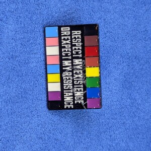 May include: A rectangular pin with a black border and a colorful design. The pin features the text "RESPECT MY EXISTENCE OR EXPECT MY RESISTANCE" in white. The design includes a series of colored blocks.