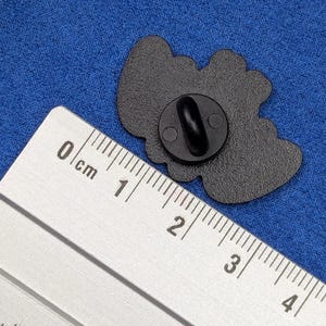 May include: Black enamel pin with a rounded shape. The pin has a black clasp on the back. A ruler is placed next to the pin for scale. The pin is on a blue fabric surface.