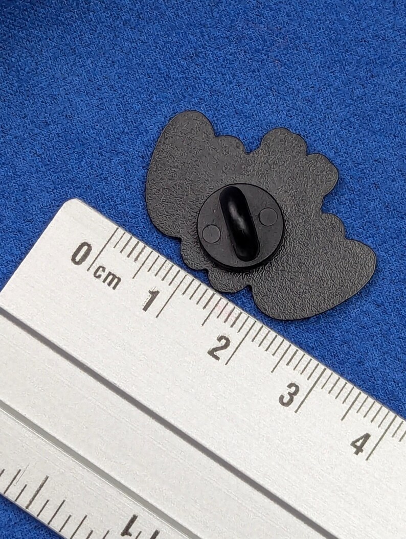 May include: Black enamel pin with a rounded shape. The pin has a black clasp on the back. A ruler is placed next to the pin for scale. The pin is on a blue fabric surface.