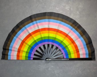 Puede incluir: Un abanico plegable con un diseño vibrante de la bandera progress pride. El abanico muestra arcos concéntricos de colores negro, marrón, azul claro, rosa, blanco, rojo, naranja, amarillo, verde, azul y morado, con una estructura de plástico negro.