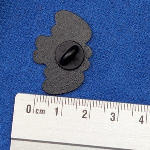 May include: A black enamel pin with a butterfly design. The pin is approximately 1 inch in length and has a black clutch back. The pin is shown next to a ruler for scale.