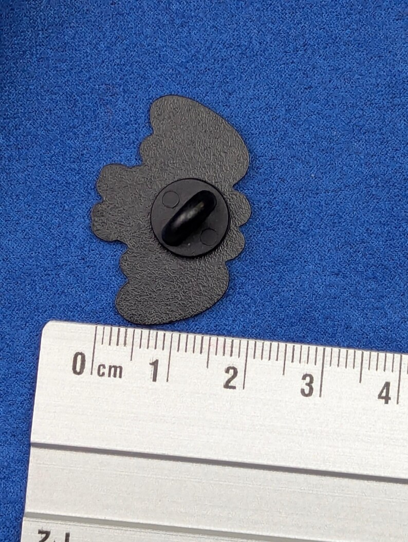 May include: A black enamel pin with a butterfly design. The pin is approximately 1 inch in length and has a black clutch back. The pin is shown next to a ruler for scale.