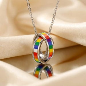 Peut inclure: Un collier argenté avec deux anneaux entrelacés, chacun orné d'un motif émaillé arc-en-ciel. Le collier est suspendu à une fine chaîne. Le motif des anneaux se reflète sur la surface en dessous.