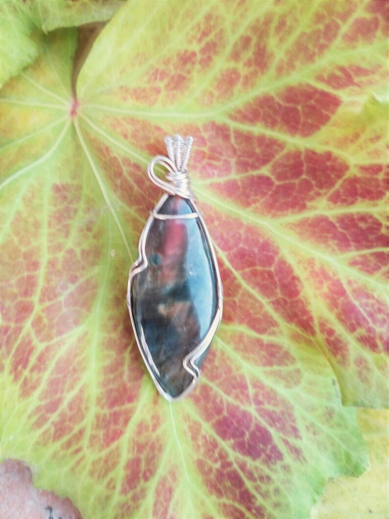 May include: A silver wire wrapped pendant featuring a dark green, brown, and red gemstone with a unique, irregular shape.