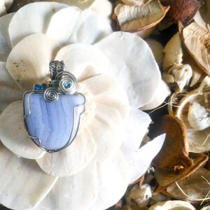 May include: A blue agate pendant with silver wire wrapping and blue crystal accents. The pendant is displayed on a white flower.