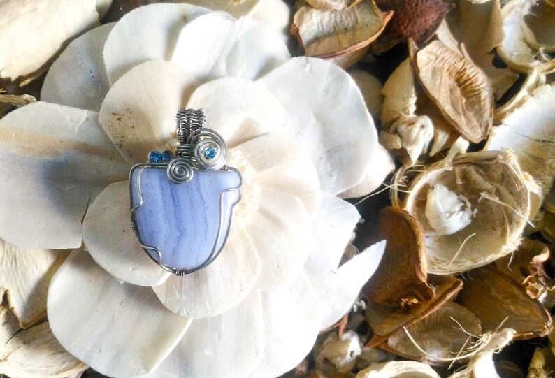 May include: A blue agate pendant with silver wire wrapping and blue crystal accents. The pendant is displayed on a white flower.