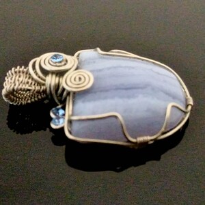 May include: A wire-wrapped pendant featuring a light blue agate stone with a swirling pattern. The stone is surrounded by silver wire and accented with two small blue crystals.