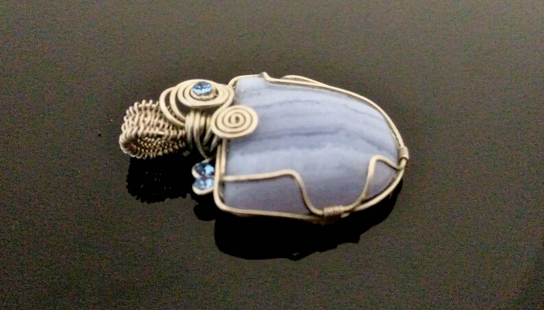 May include: A wire-wrapped pendant featuring a light blue agate stone with a swirling pattern. The stone is surrounded by silver wire and accented with two small blue crystals.