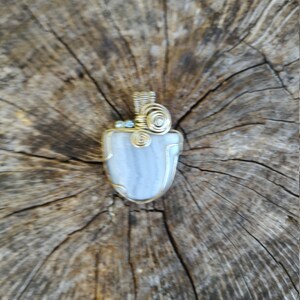 May include: A silver and gold wire wrapped pendant featuring a light blue agate stone. The pendant is on a wooden background.