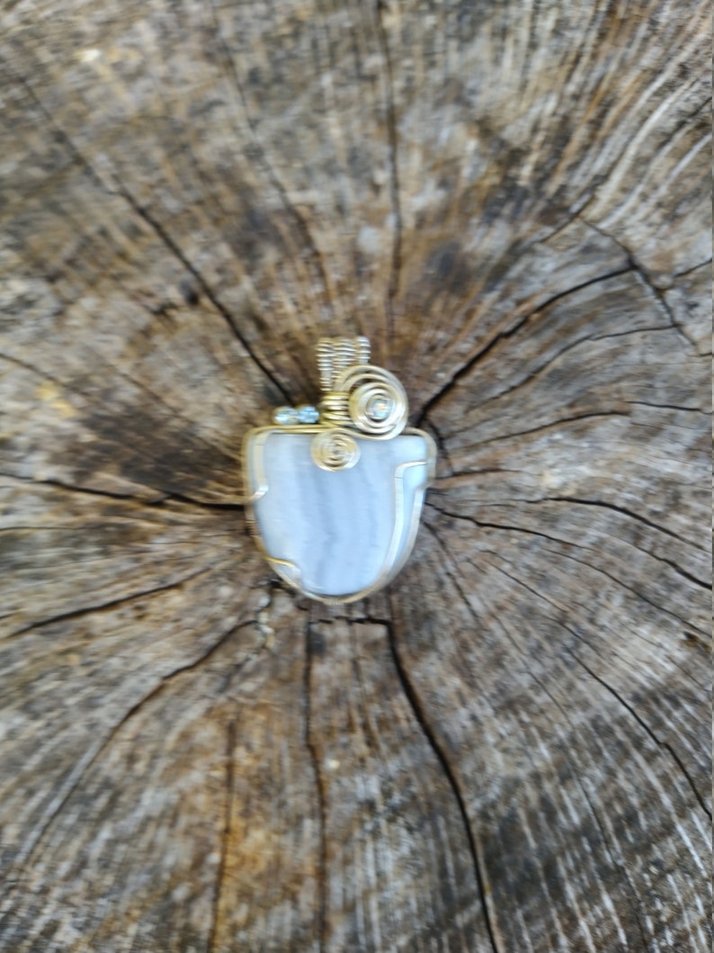 May include: A silver and gold wire wrapped pendant featuring a light blue agate stone. The pendant is on a wooden background.