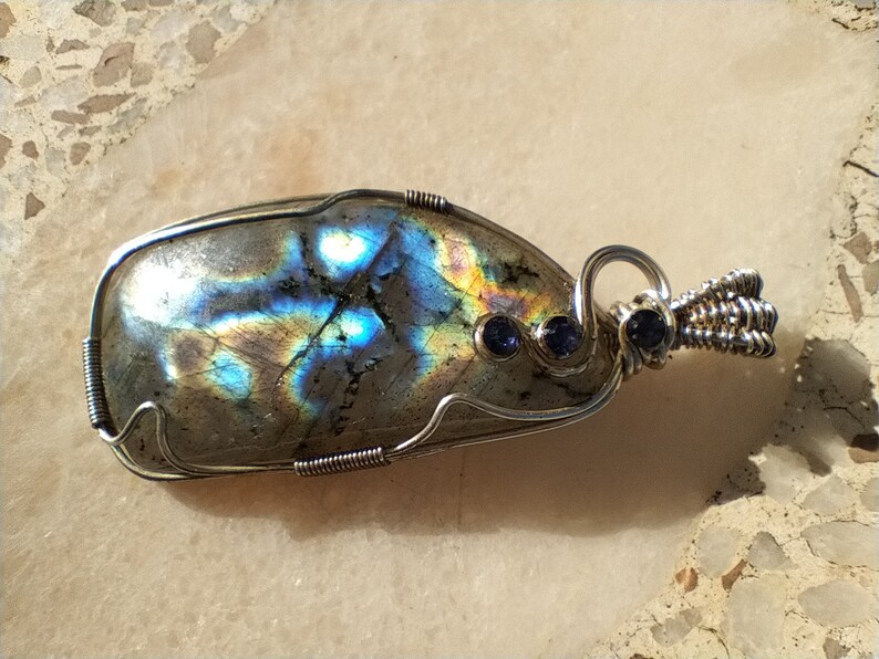 May include: A silver wire wrapped pendant featuring a large iridescent labradorite gemstone with two smaller blue sapphire accents.