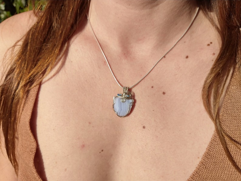 May include: A silver chain necklace with a pendant featuring a light blue agate stone wrapped in silver wire and accented with small blue and clear gemstones.