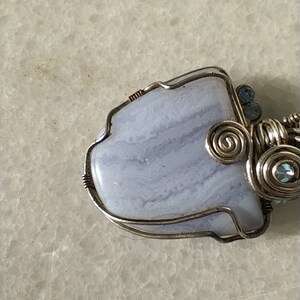 May include: A silver wire wrapped pendant featuring a light blue agate stone with white and gray swirls. The pendant is accented with small silver and blue beads.