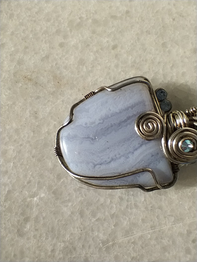 May include: A silver wire wrapped pendant featuring a light blue agate stone with white and gray swirls. The pendant is accented with small silver and blue beads.