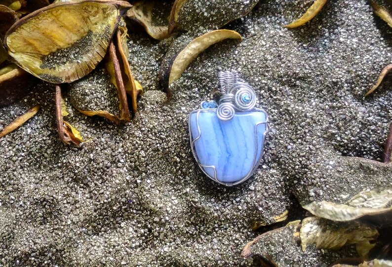 May include: A blue agate pendant wrapped in silver wire with blue crystal accents. The pendant is sitting on a bed of silver glitter.