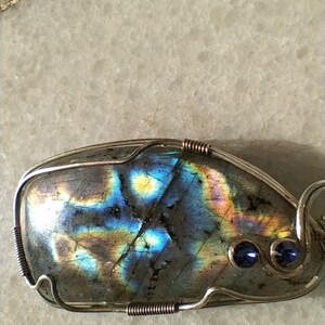 May include: A silver wire wrapped pendant featuring a large iridescent labradorite gemstone with blue accents. The pendant has a unique, organic shape and is set with two small blue gemstones.