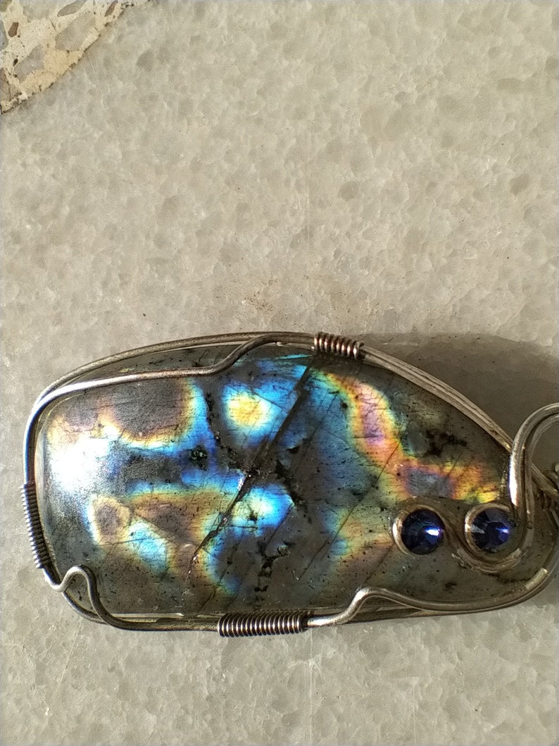 May include: A silver wire wrapped pendant featuring a large iridescent labradorite gemstone with blue accents. The pendant has a unique, organic shape and is set with two small blue gemstones.