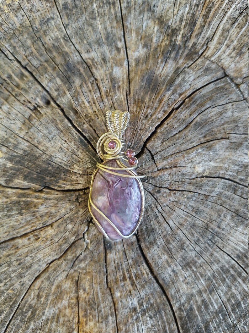 May include: A purple and silver wire-wrapped pendant featuring a purple gemstone with a swirling pattern. The pendant is accented with two small pink gemstones.