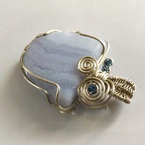 May include: A silver wire wrapped pendant featuring a light blue agate stone with swirls of white and a silver wire design with blue rhinestones.