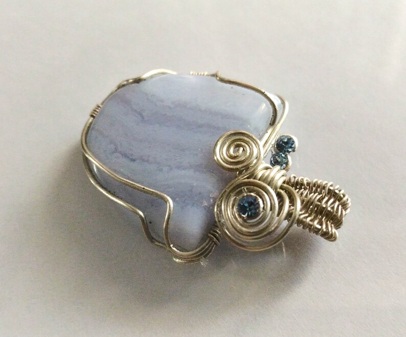 May include: A silver wire wrapped pendant featuring a light blue agate stone with swirls of white and a silver wire design with blue rhinestones.