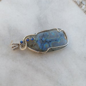 May include: A silver wire wrapped pendant featuring a blue-grey labradorite gemstone with iridescent blue and green hues. Three small blue gemstones are set in the wire wrapping.