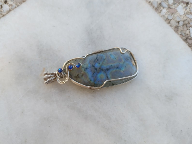 May include: A silver wire wrapped pendant featuring a blue-grey labradorite gemstone with iridescent blue and green hues. Three small blue gemstones are set in the wire wrapping.