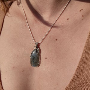 May include: A silver chain necklace with a large, grey, irregularly shaped stone pendant. The stone is set in silver wire and has black accents.