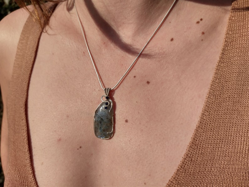 May include: A silver chain necklace with a large, grey, irregularly shaped stone pendant. The stone is set in silver wire and has black accents.