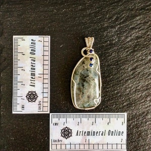 May include: A silver wire wrapped pendant featuring a gray and green labradorite stone with two small blue gemstones. The pendant is approximately 2 inches in length.