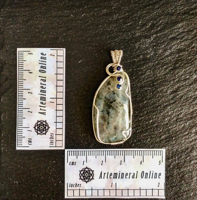 May include: A silver wire wrapped pendant featuring a gray and green labradorite stone with two small blue gemstones. The pendant is approximately 2 inches in length.