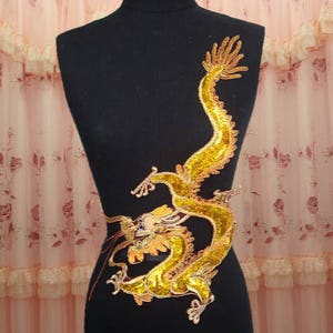 Könnte beinhalten: Goldene Pailletten-Drachen-Applikation mit detailliertem Design. Der Drache blickt nach links und hat einen langen Schwanz. Die Applikation ist perfekt, um jedem Projekt einen Hauch von Glanz und Verspieltheit zu verleihen.