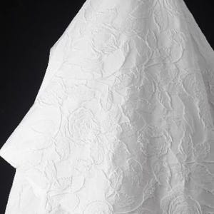 Puede incluir: Tela blanca con un patrón floral en relieve. La tela está doblada en forma de triángulo.