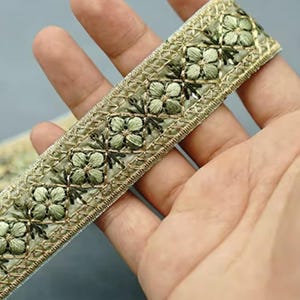 May include: Close-up of a decorative trim featuring a floral pattern. The trim has a light green and gold color scheme, with embroidered flowers and a metallic gold border. The trim is held in a hand.