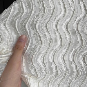 Puede incluir: Tela blanca con un patrón ondulado y texturado. La tela es suave y tiene un brillo sutil.