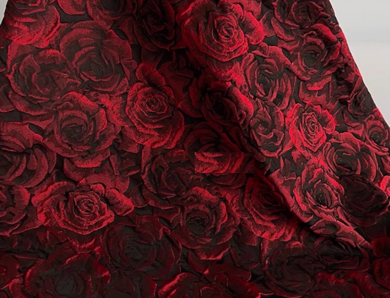 May include: Red fabric with a raised rose pattern. The roses are a deep red colour and are arranged in a repeating pattern.