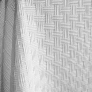 May include: Close-up of a white woven fabric with a textured basketweave pattern. The fabric is draped over an unseen object, creating soft folds and shadows. The material appears to be thick and durable, suitable for home decor.