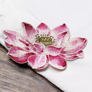 May include: A pink and white embroidered lotus flower patch. The flower has a detailed design with a yellow center.