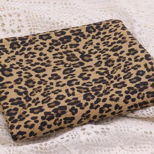 May include: A folded piece of fabric featuring a tan background with a repeating black and dark brown leopard print pattern. The textile has a visible woven texture and is arranged in a rectangular shape with clean, straight edges.