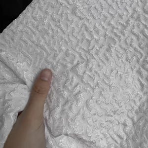 Könnte beinhalten: Nahaufnahme eines weißen Stoffes mit einem erhabenen, strukturierten Muster. Der Stoff hat ein gewebtes Aussehen mit einem sich wiederholenden Design. Das Material ist weich und scheint gefaltet zu sein, mit einer Hand, die den Stoff berührt.