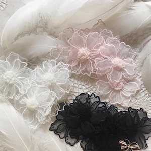 May include: Close-up of floral lace appliques in white, pale pink, and black. The delicate flowers have a layered petal design and a central, textured core. White feathers and lace fabric provide a soft, textured background.