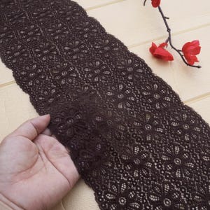 May include: Dark brown lace fabric with a floral pattern. The lace is held by a hand, showcasing the intricate design. The fabric is a rich, deep brown color, and the floral pattern is detailed and delicate. The lace is likely used for sewing, crafts, or apparel.