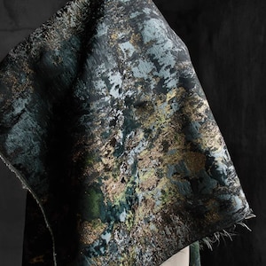 May include: A close-up of a dark green fabric with a metallic gold and silver abstract pattern. The fabric has a textured surface and a slightly frayed edge.
