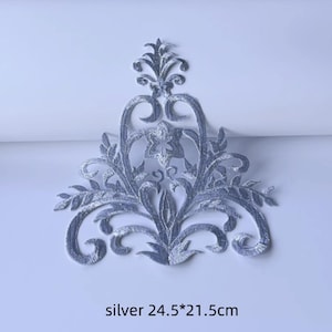May include: Silver embroidered applique with an ornate, symmetrical design. The applique features scrolling patterns, floral elements, and a central star-shaped detail. The text "silver 24.5*21.5cm" is at the bottom. The applique measures approximately 9.6 x 8.5 inches.