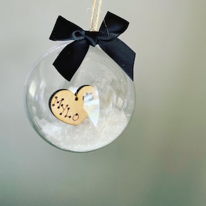 Puede incluir: Un adorno de vidrio transparente con una cinta negra y un corazón de madera con el nombre "MYLO" inscrito en él. El adorno está lleno de purpurina blanca.