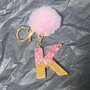 May include: A pink and gold glitter letter K keychain with a pink fluffy pom-pom charm.