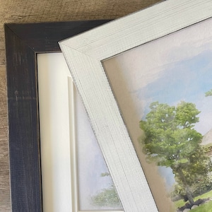 May include: Two picture frames, one black and one white, are shown side-by-side. The white frame is slightly angled and shows a watercolor painting of a house with trees and a blue sky.