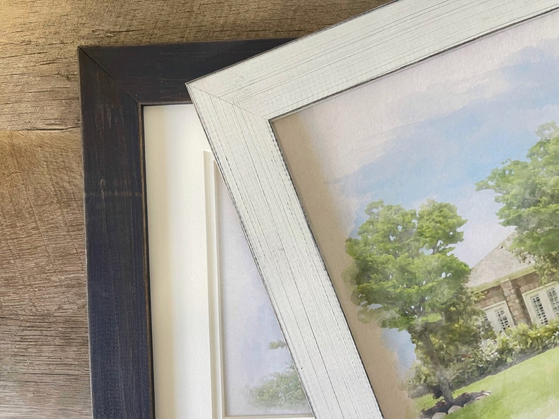 May include: Two picture frames, one black and one white, are shown side-by-side. The white frame is slightly angled and shows a watercolor painting of a house with trees and a blue sky.