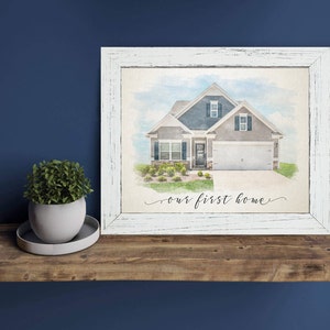 May include: A watercolor painting of a house with a gray exterior and a white trim. The house has a blue roof and a white door. The painting is framed in a white frame with a distressed finish. The text "our first home" is written in black cursive below the house.