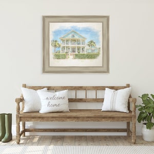 May include: A watercolor painting of a light blue house with white trim and a white porch. The house is surrounded by green bushes and trees. The painting is framed in a light brown frame and is hanging on a white wall. A wooden bench with white pillows and a green plant in a white pot are in the foreground.